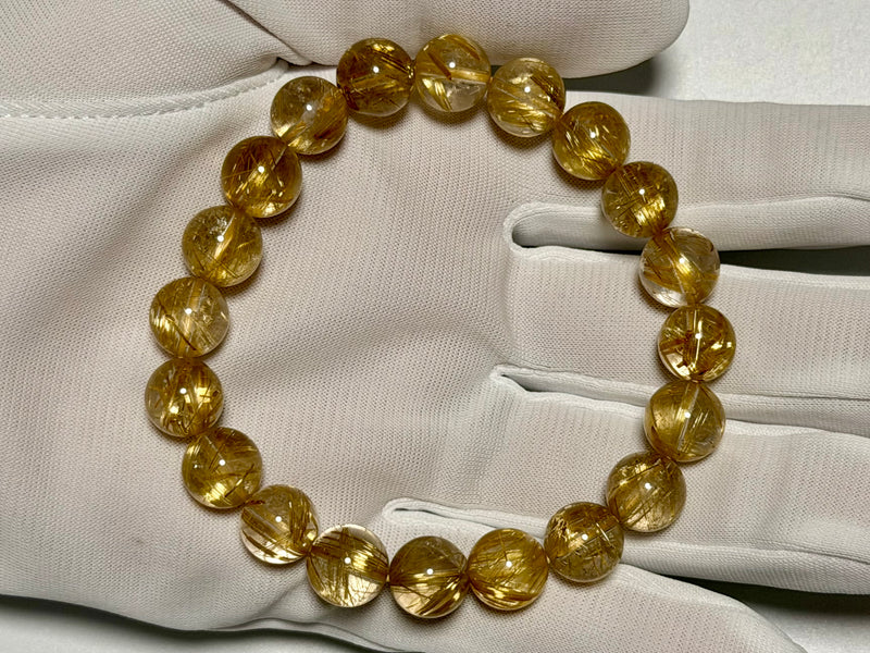 Clear Rutilated Quartz bracelet, 10–10.5 mm, gloved-hand view showing fine gold rutile needles and quartz translucency.
