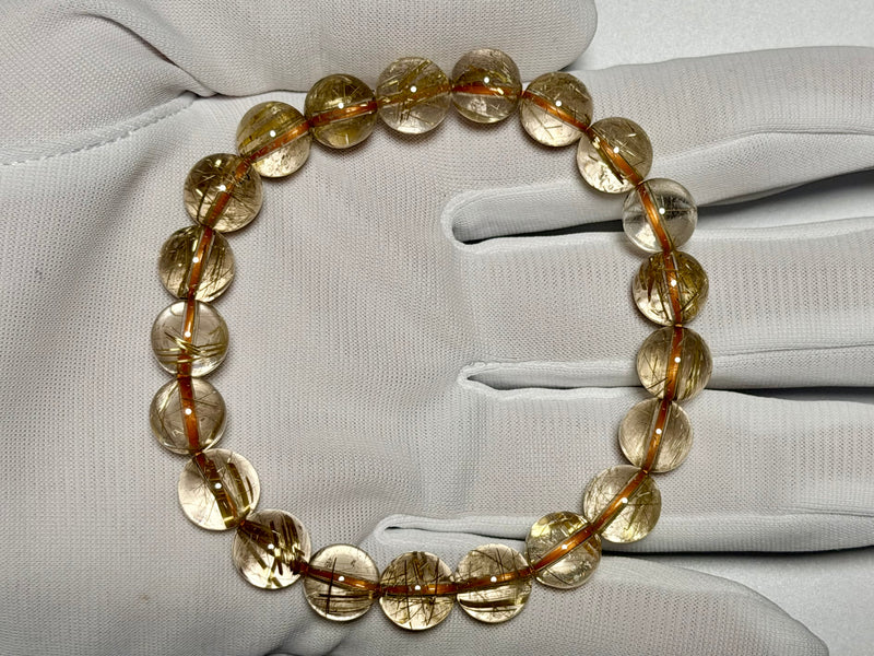 Clear Rutilated Quartz bracelet, 9.5 mm, gloved-hand view showing fine gold rutile needles and quartz translucency.